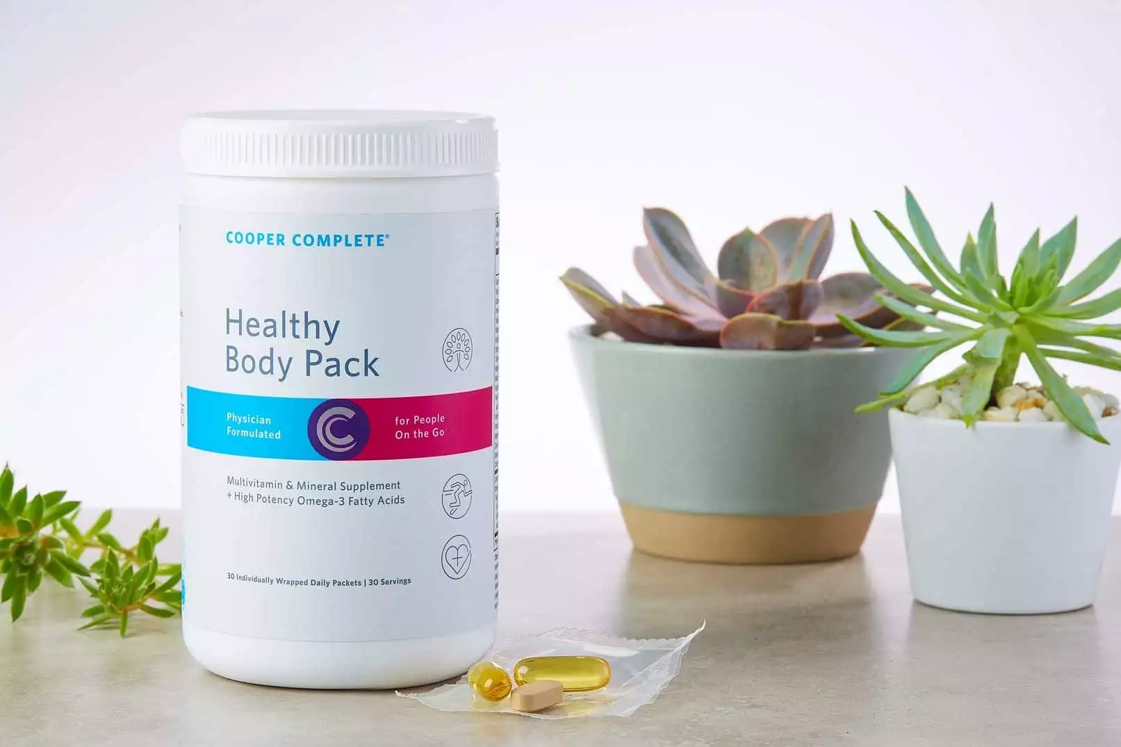 Cooper Complete Healthy Body Daily Vitamin Pack dietary supplement canister and a single daily packet on a counter with several succulent plants. Daily Vitamin Pack for adults; daily vitamin pack for women; daily vitamin pack for men