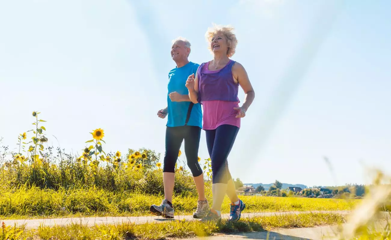 Photo of a mature man and woman in athletic wear walking on a park path past sunflowers.