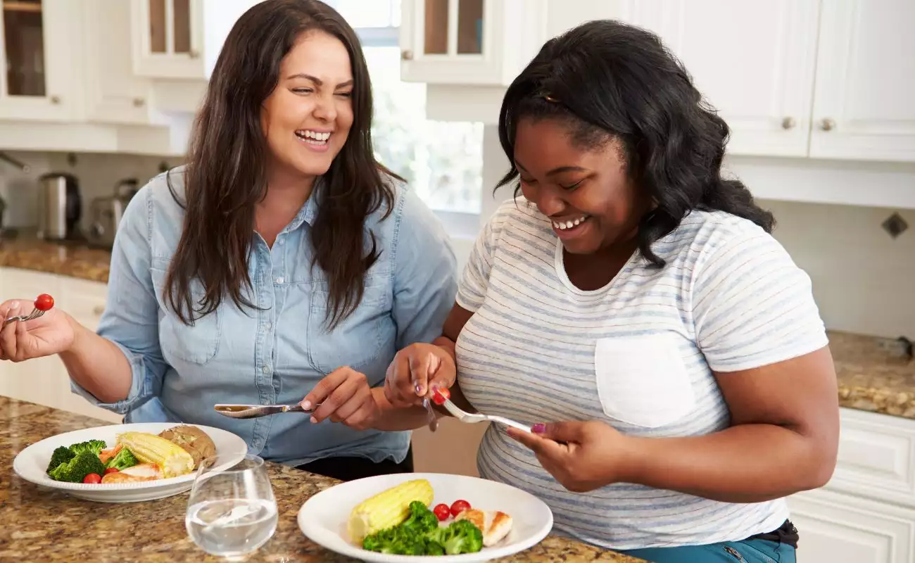 Photo of two women in the kitchen enjoying a laugh and eating a meal.